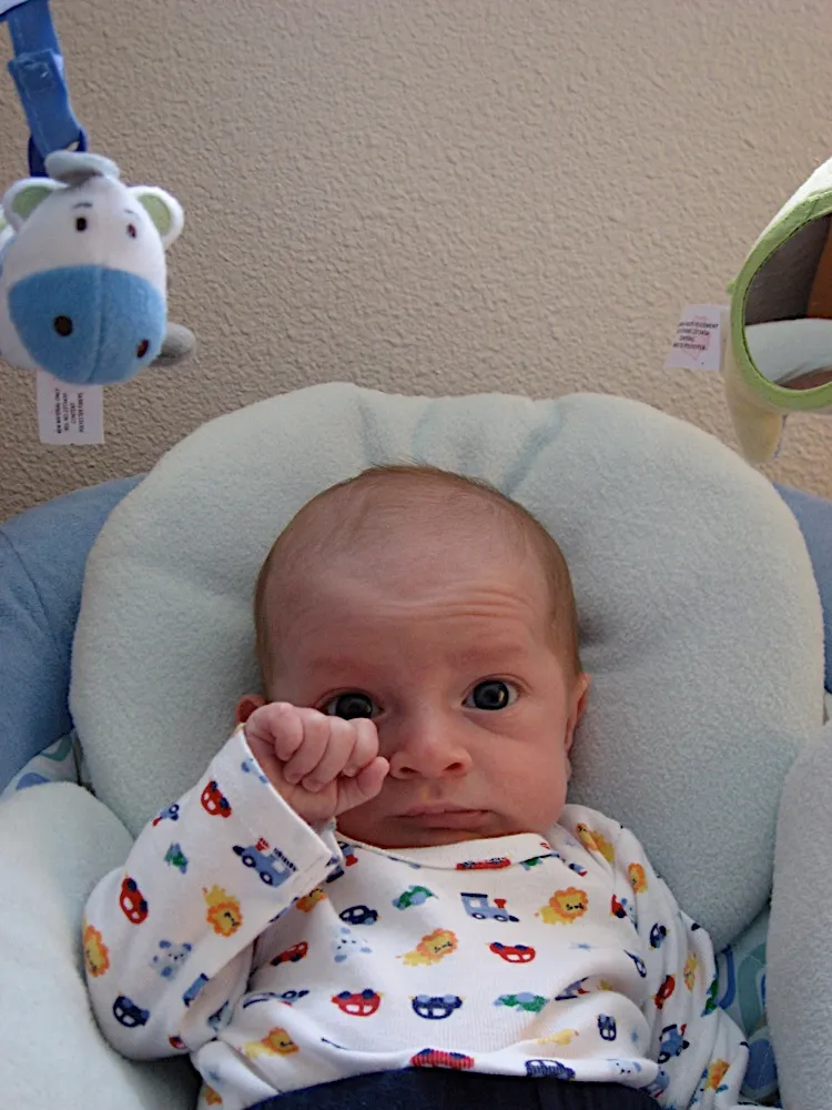 Rubbing one eye with a fist, eyebrows raised, sitting in the bouncer seat.
