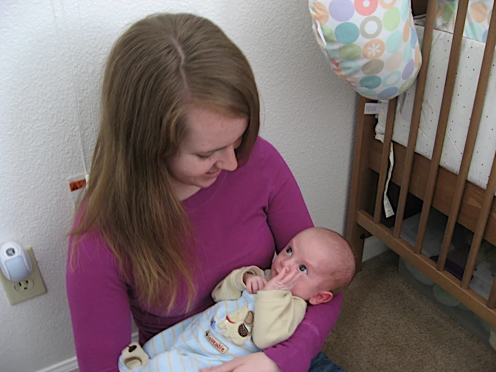 A woman smiles down at him while holding him near his crib, his hand near his mouth.