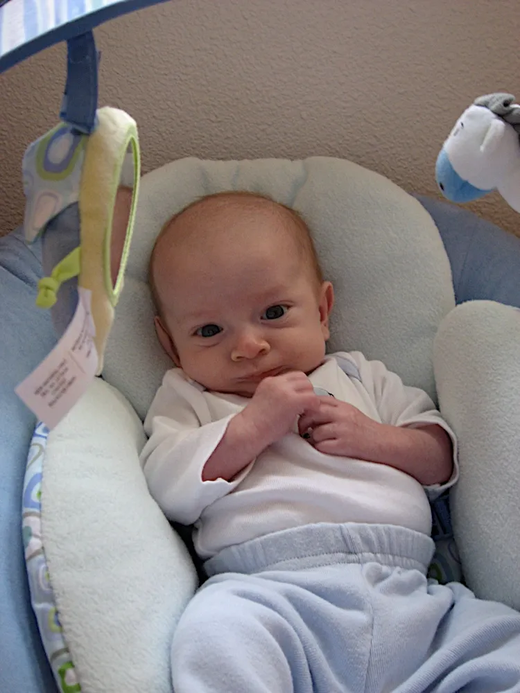 In the bouncer seat with hands clasped near his chin, wearing white and pale blue.