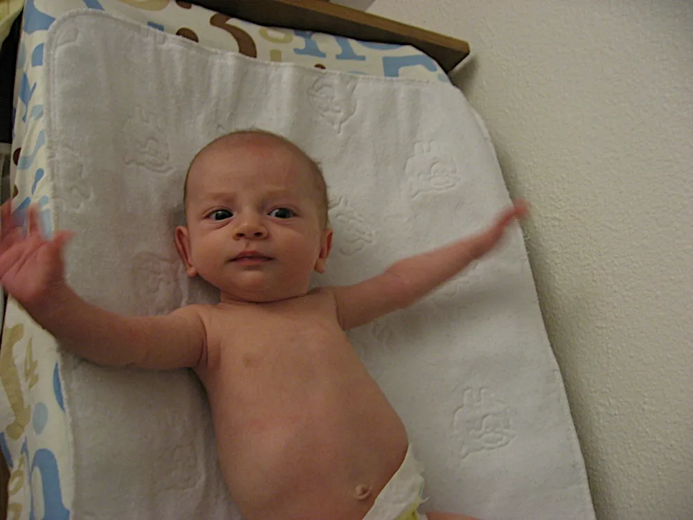 Shirtless on a changing pad with both arms flung out wide, looking alert.