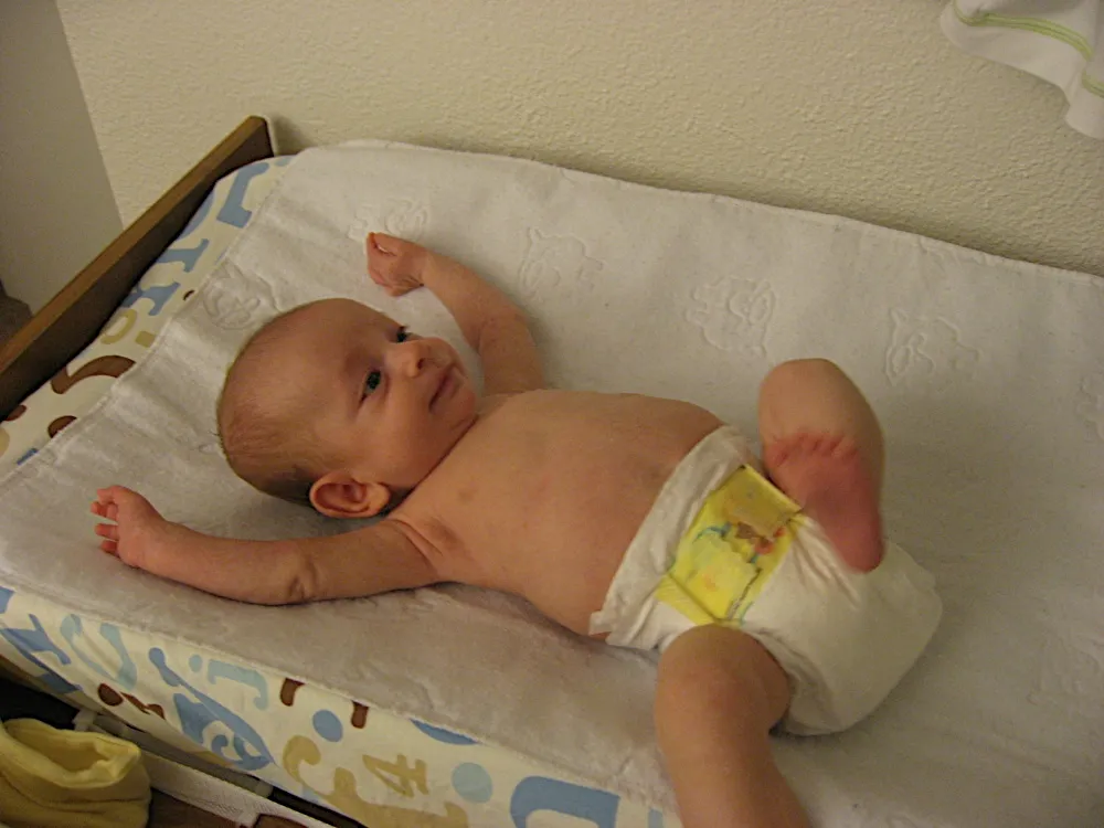 In just a diaper on the changing pad, one knee bent and one arm raised, smiling.