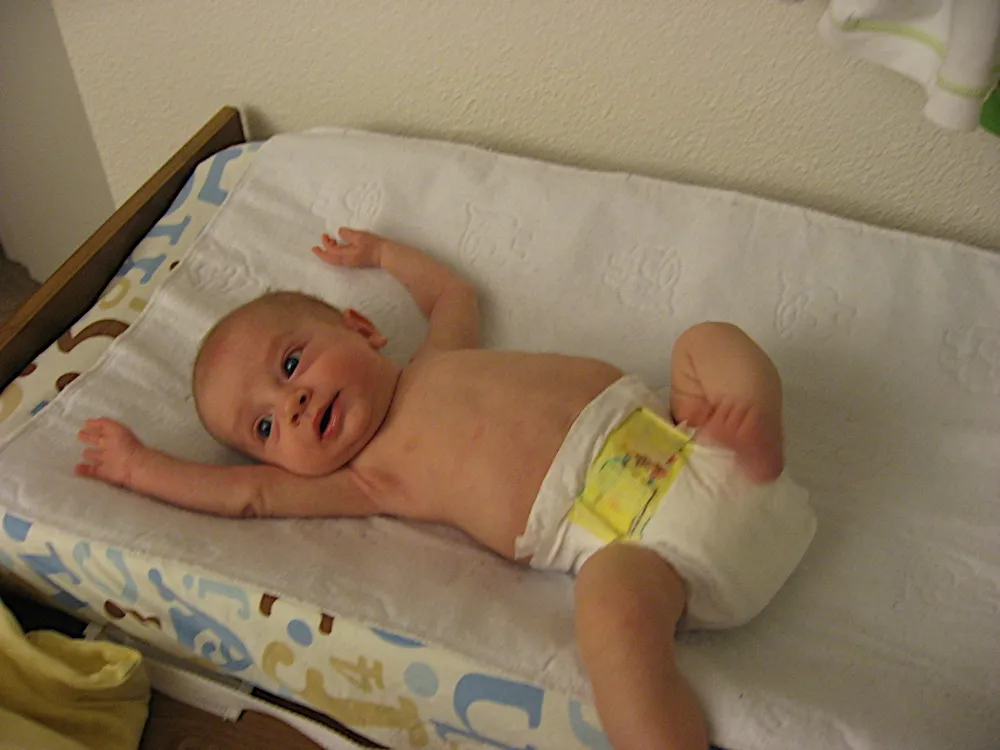 On the changing pad in just a diaper, mouth open, looking toward the camera.