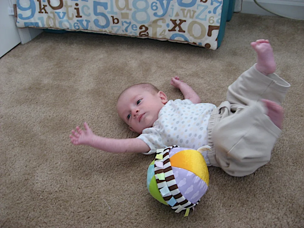 On the carpet with legs kicked up, a colorful soft ball toy resting on his stomach.
