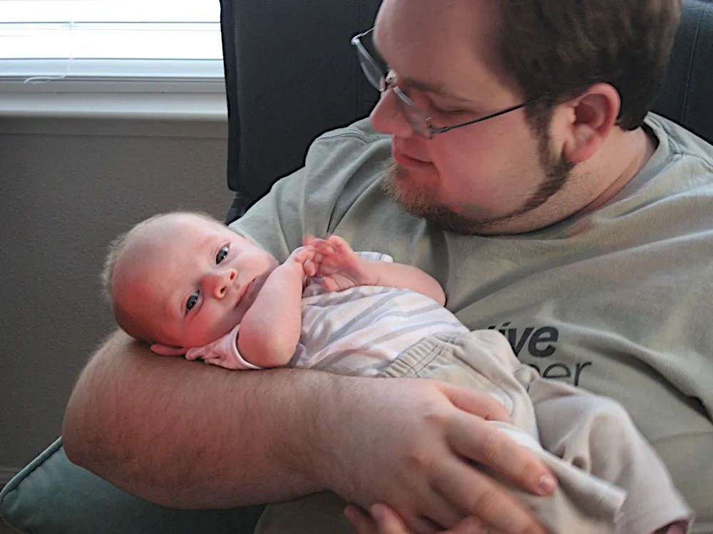 Resting in the crook of a bearded man's arm, looking at the camera while he smiles down at him.