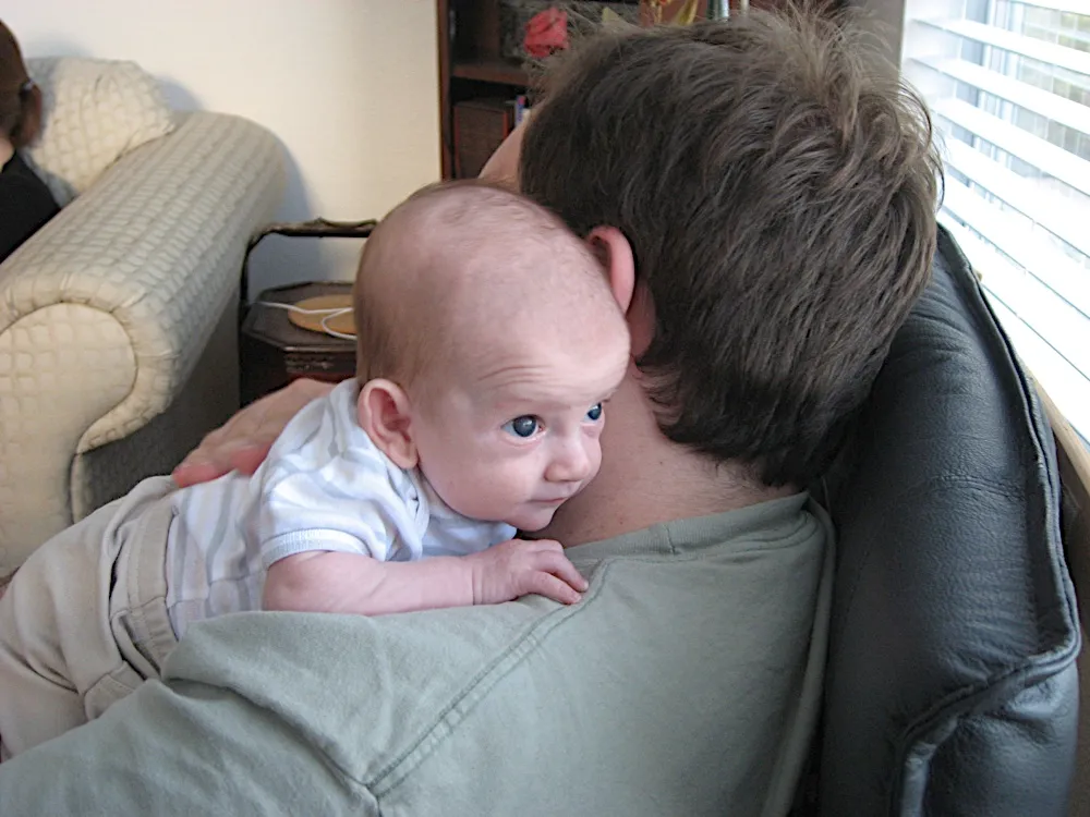 Held upright over a man's shoulder, chin resting there, peering at the camera.