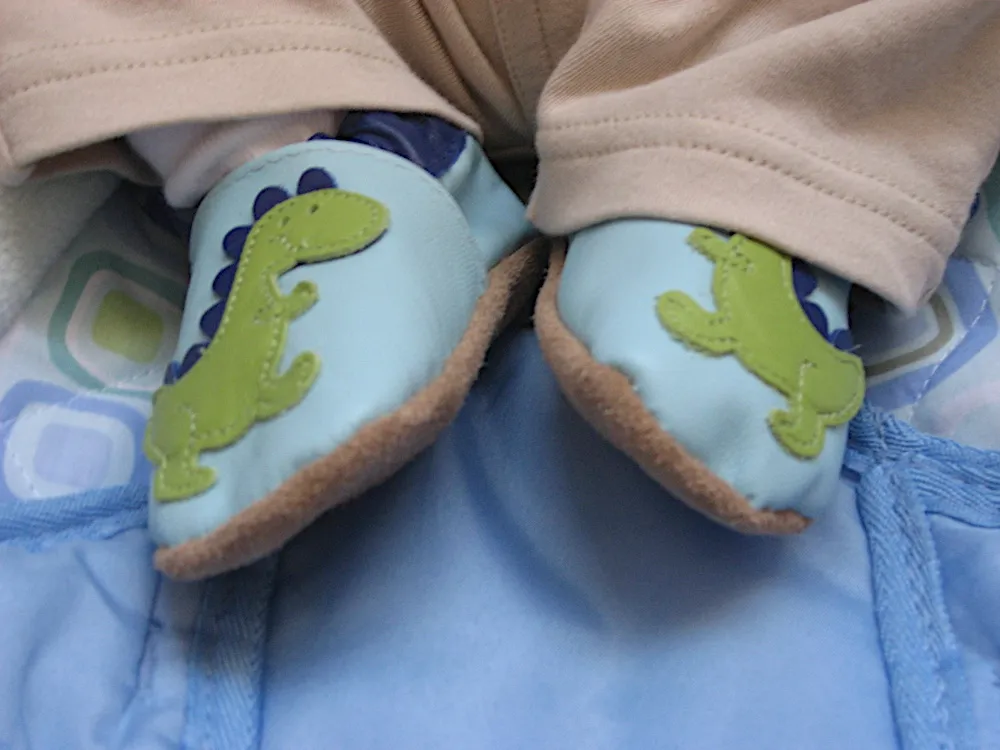 Close-up of soft-soled baby shoes with green dinosaur appliqués, feet tucked into tan pants.