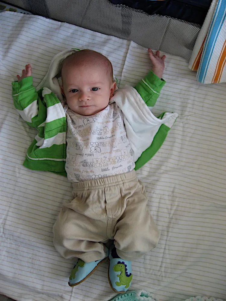Laid out in a playpen wearing the green striped hoodie and dinosaur shoes, arms raised.