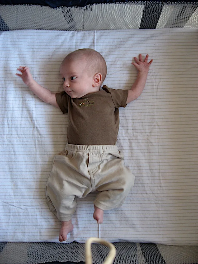 Lying in a playpen with both arms raised, wearing a brown shirt, looking off to the side.