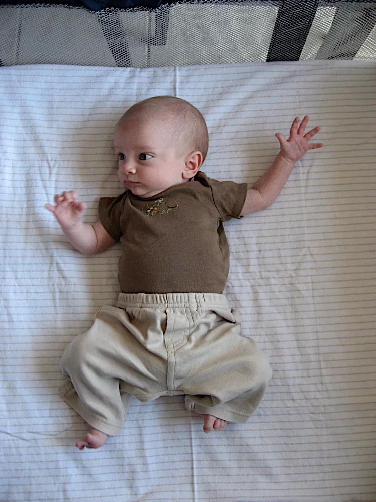 Knees drawn up in a playpen, wearing a brown shirt, glancing toward his own hand.
