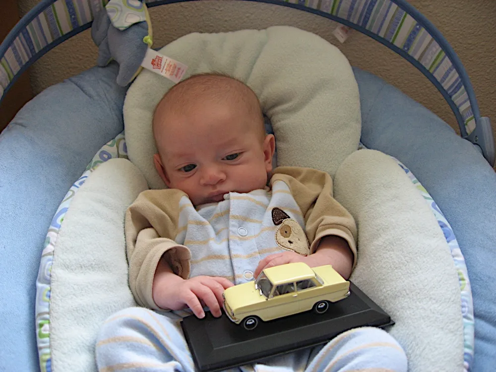 In the bouncer seat with one hand resting on a small yellow model car.